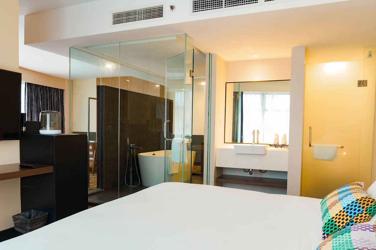 A modern hotel room with a large bed, a glass-enclosed shower area, a vanity with a mirror, and a television on a stand.