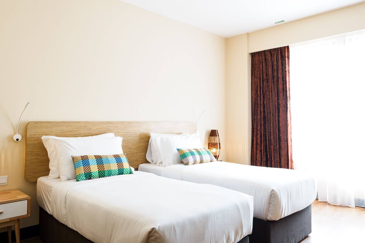 A hotel room with two beds, a bedside table, a lamp, and a large window with curtains.