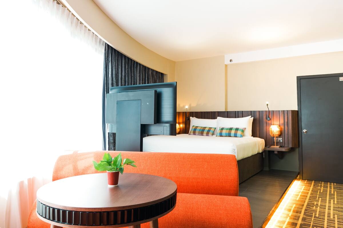 A hotel room with a bed, orange seating, a small table with a plant, a TV, and a bedside lamp.