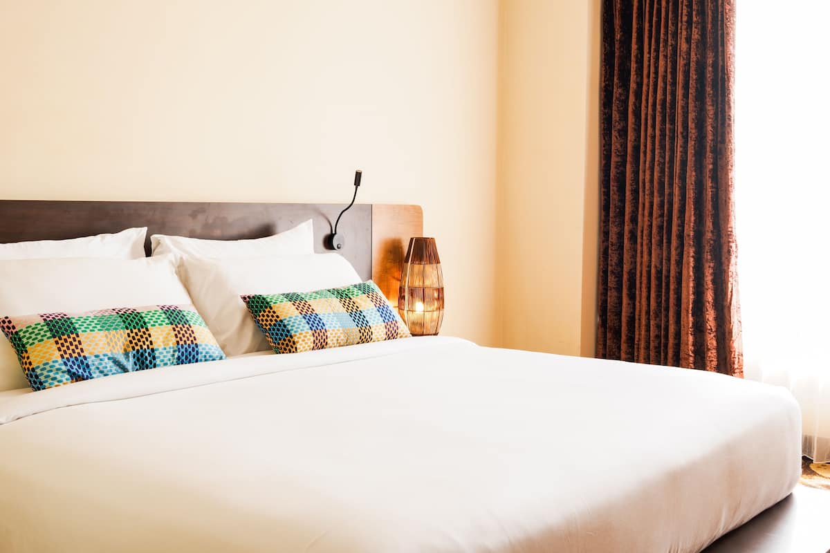 A neatly made bed with white linens and two patterned pillows, a bedside table with a lamp, and a window with curtains.