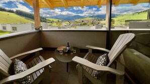 Un balcon avec des poutres en bois, deux chaises, une petite table et une vue sur un paysage verdoyant parsemé de bâtiments.