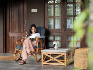 A person sitting on a wicker chair reading a magazine, with a wooden table and a vase of flowers nearby. - Club Mango (Kandakuliya)