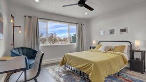 Un dormitorio con cama, escritorio, silla, ventilador de techo y ventana con cortinas.