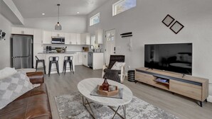 Una cocina moderna con armarios blancos, un microondas y una isla grande. Hay un comedor con una mesa y sillas, un sofá de cuero y un televisor sobre un soporte de madera.