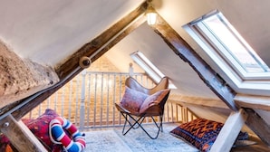 Ein Schlafzimmer im Dachgeschoss mit schrägen Wänden, einem Holzstuhl, einem farbenfrohen Kissen und einem kleinen Fenster.