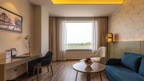 A hotel room with a desk, two chairs, a sofa, a round table with a fruit bowl, a painting on the wall, and a large window with curtains. - Fortune Park, Hoshiarpur - Member ITC Hotels' Group (Hoshiarpur)