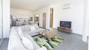 A modern living room with a sofa, coffee table, and TV. There is a kitchen in the background with cabinets and a refrigerator. - Protaras Vie Bleu Villa Vb15 (Paralimni)
