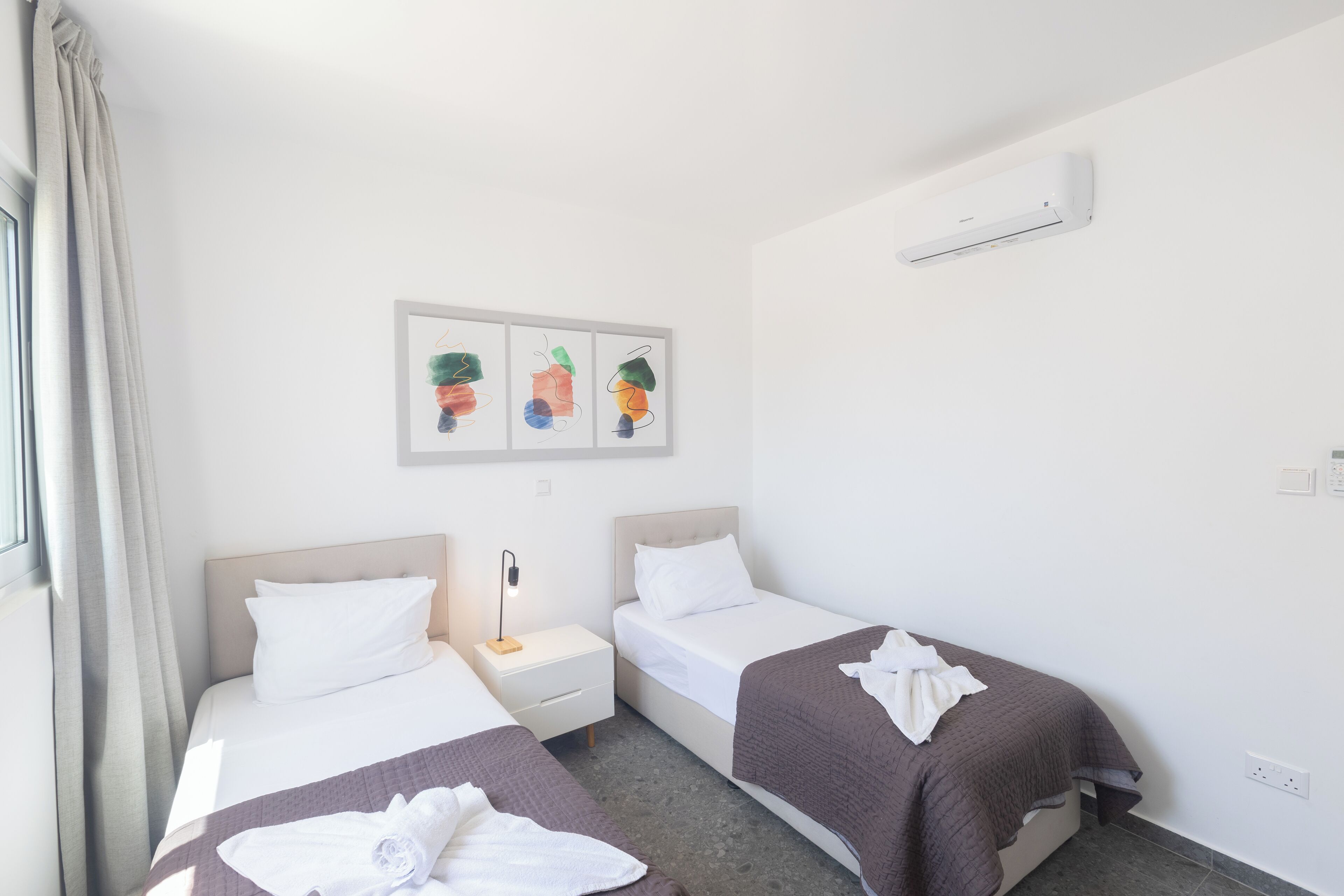 Ein Hotelzimmer mit zwei Betten, einem Nachttisch, einer Wand-Klimaanlage und einem Vorhangfenster.