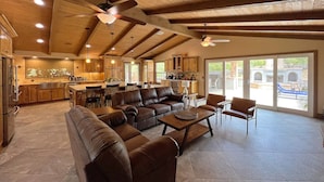 A spacious living area with a large brown leather sofa, a coffee table, and a dining table. There is a kitchen area with wooden cabinets and a dining table. The space has a high ceiling with exposed beams and a large window providing a view of the outdoors. - Villa 5B/3B with pool/spa/mini golf putting (Temecula)