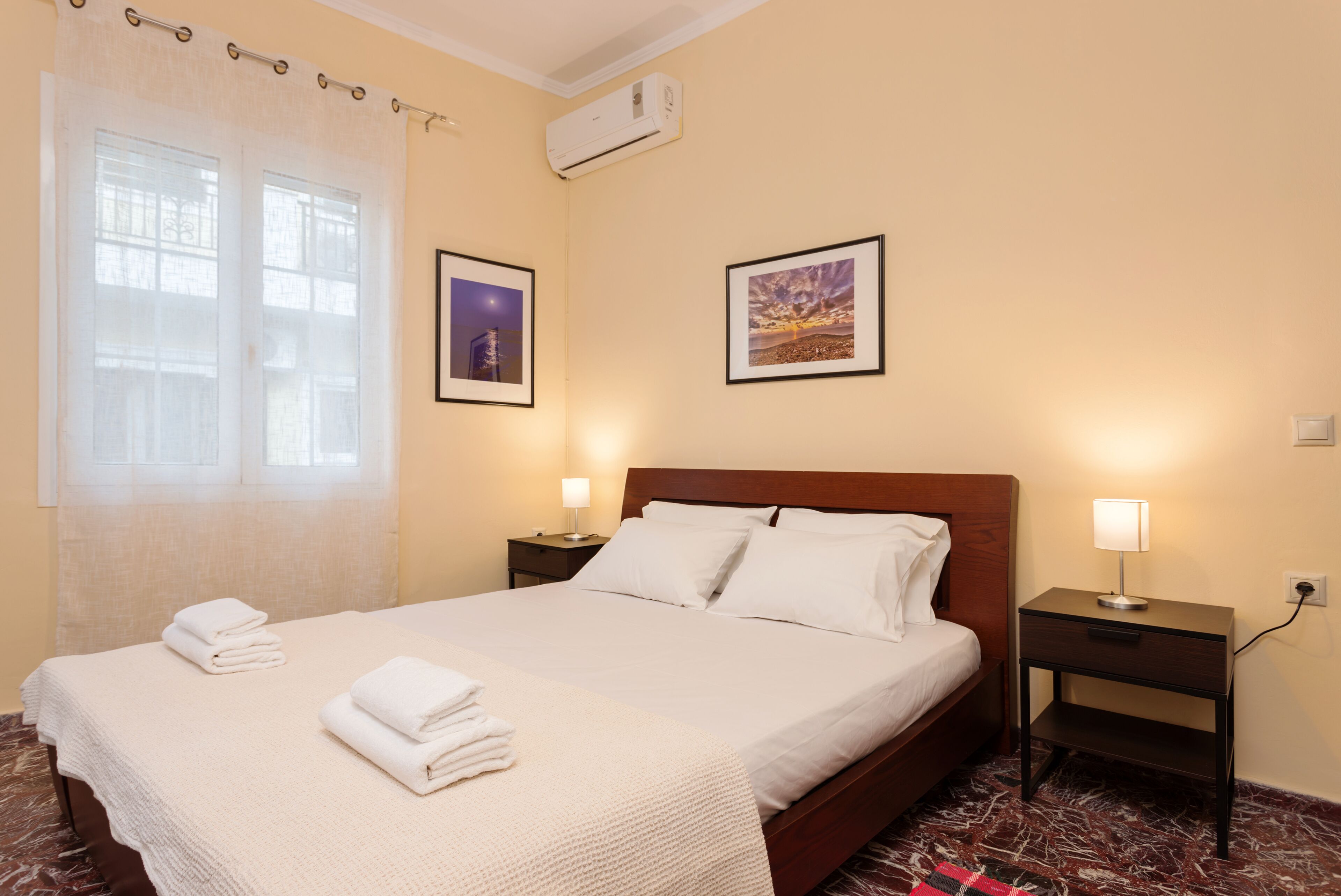 Una habitación de hotel con una cama, dos mesitas de noche con lámparas, un ventanal con cortinas y dos cuadros en la pared.