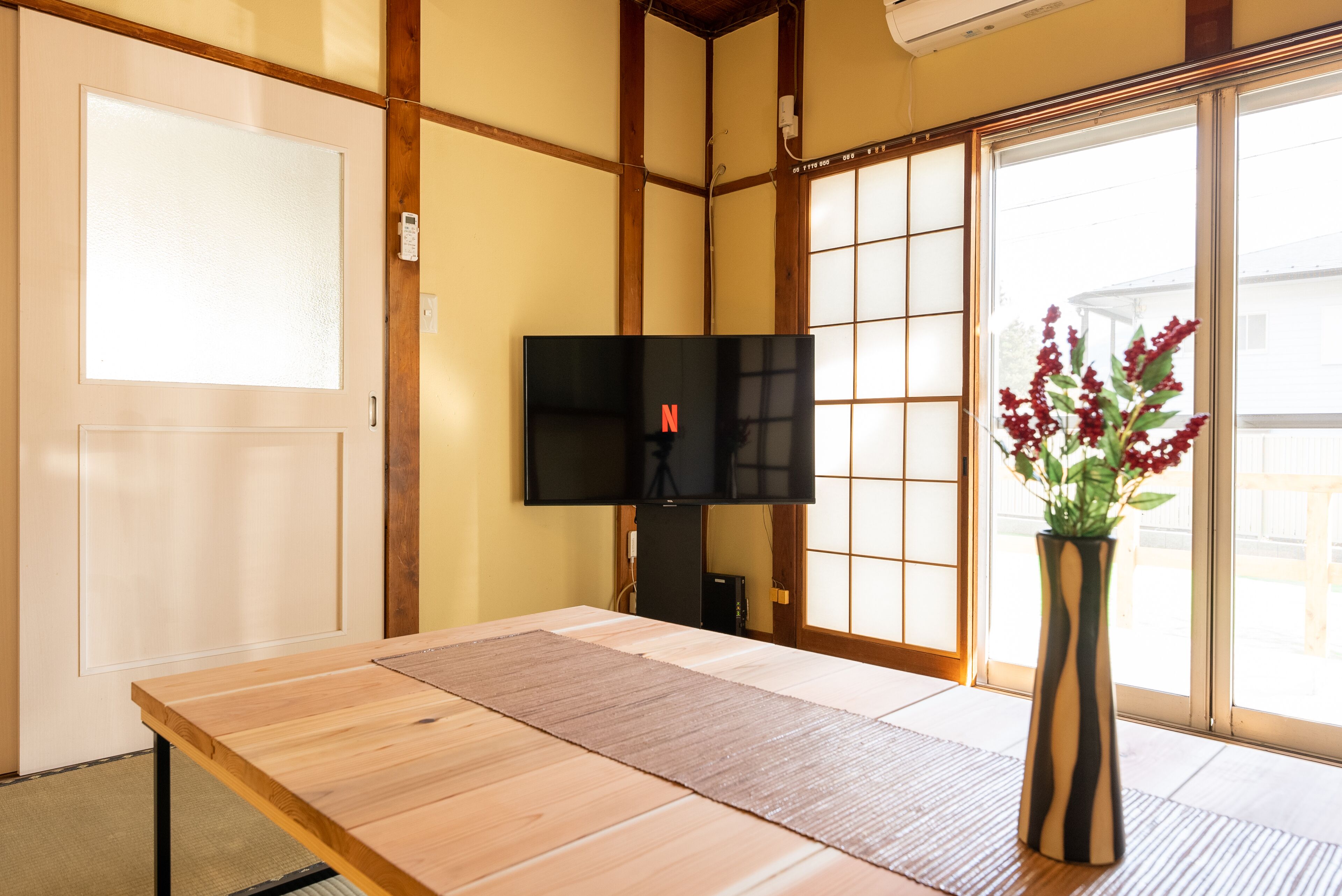 A room with a tatami mat floor, a television, a vase with flowers, and a sliding door with a view outside.