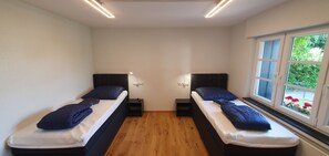 Een kamer met twee eenpersoonsbedden, houten vloeren, witte wanden en een raam met uitzicht op groen.