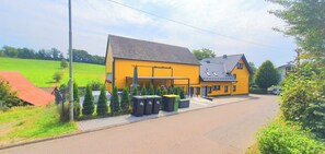 Een geel huis met een schuin dak, een omheind erf en een geplaveid pad.