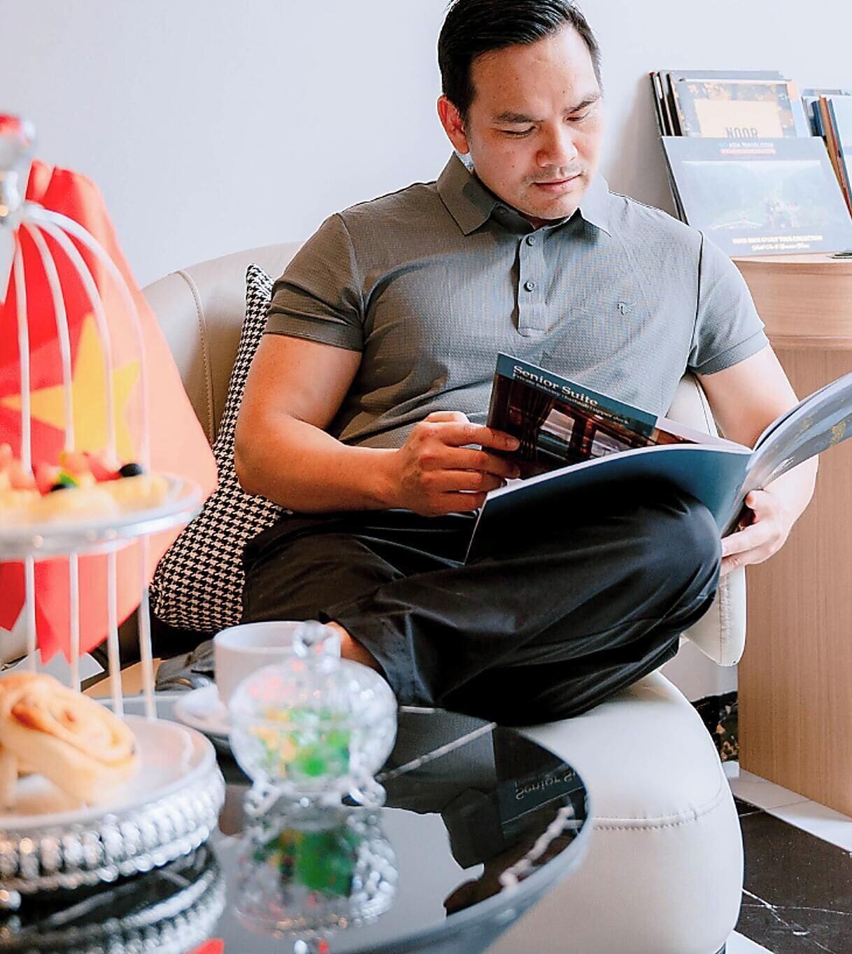 Une personne est assise sur un canapé blanc et lit un livre. Devant le canapé, il y a une table sur laquelle se trouvent un vase en verre et une assiette de nourriture.
