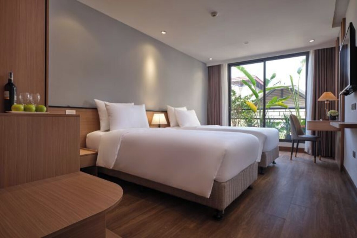 A modern hotel room with a large bed, bedside tables, a desk, and a view of greenery through the window.