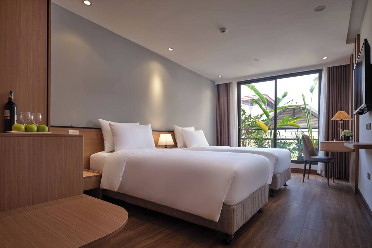 A modern hotel room with two beds, a desk, and a view of the outdoors.
