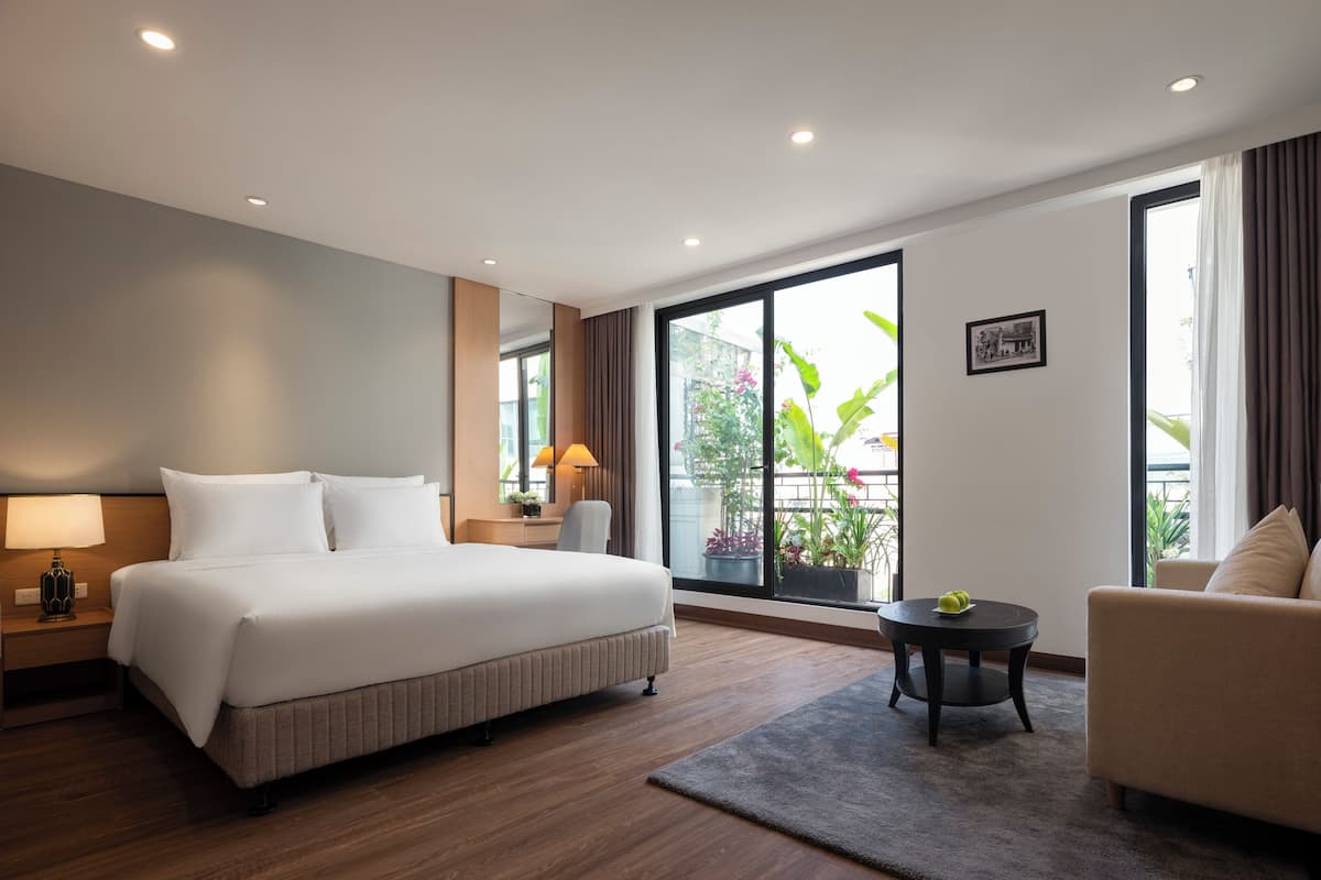 A modern hotel room with a large bed, a desk, a chair, a small table, and a sofa. There is a window with a view of the outdoors.