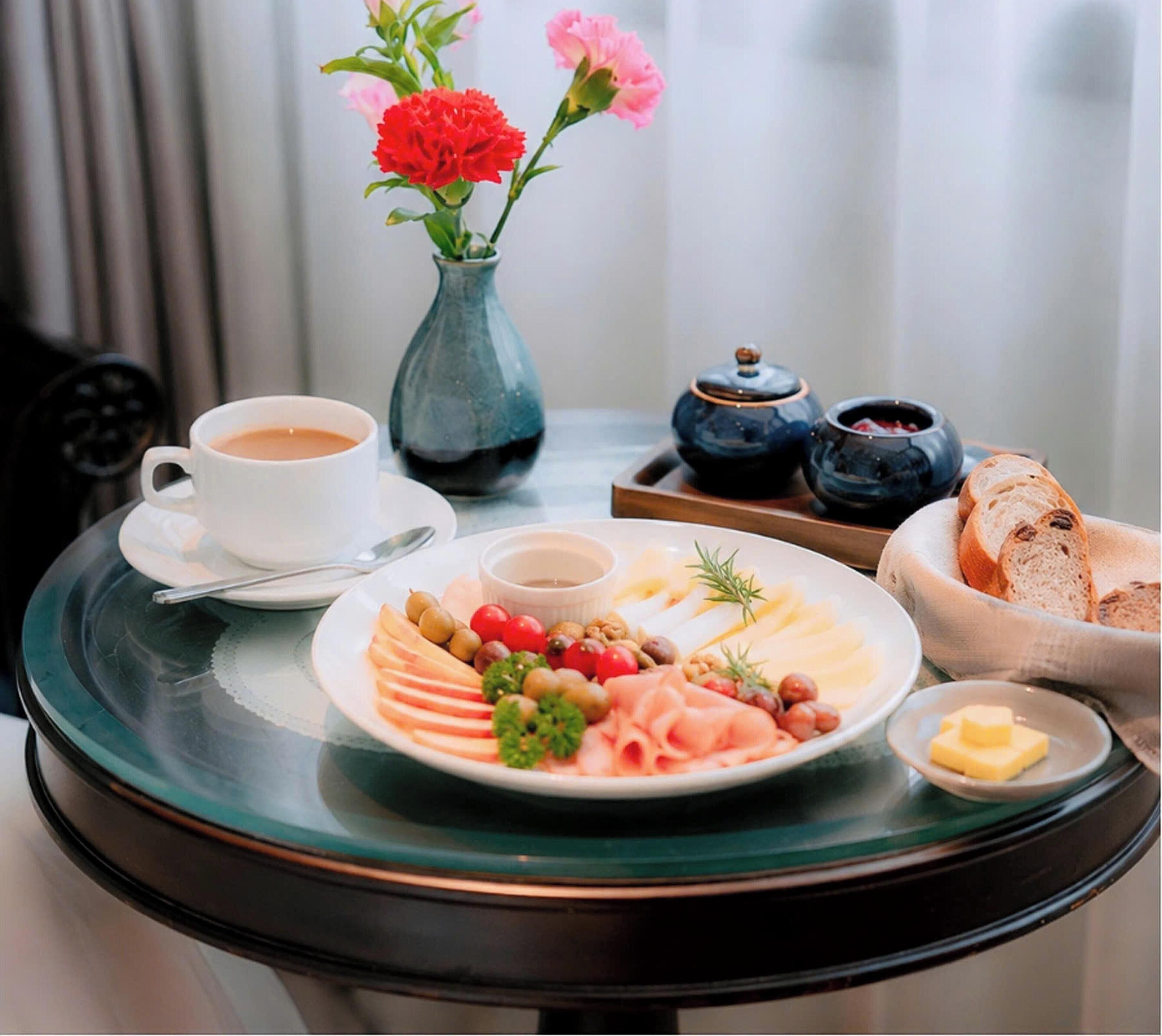 Une table mise pour le petit-déjeuner avec une assiette de nourriture, une tasse de café, un vase de fleurs et une petite table sur laquelle se trouvent du pain et des condiments.