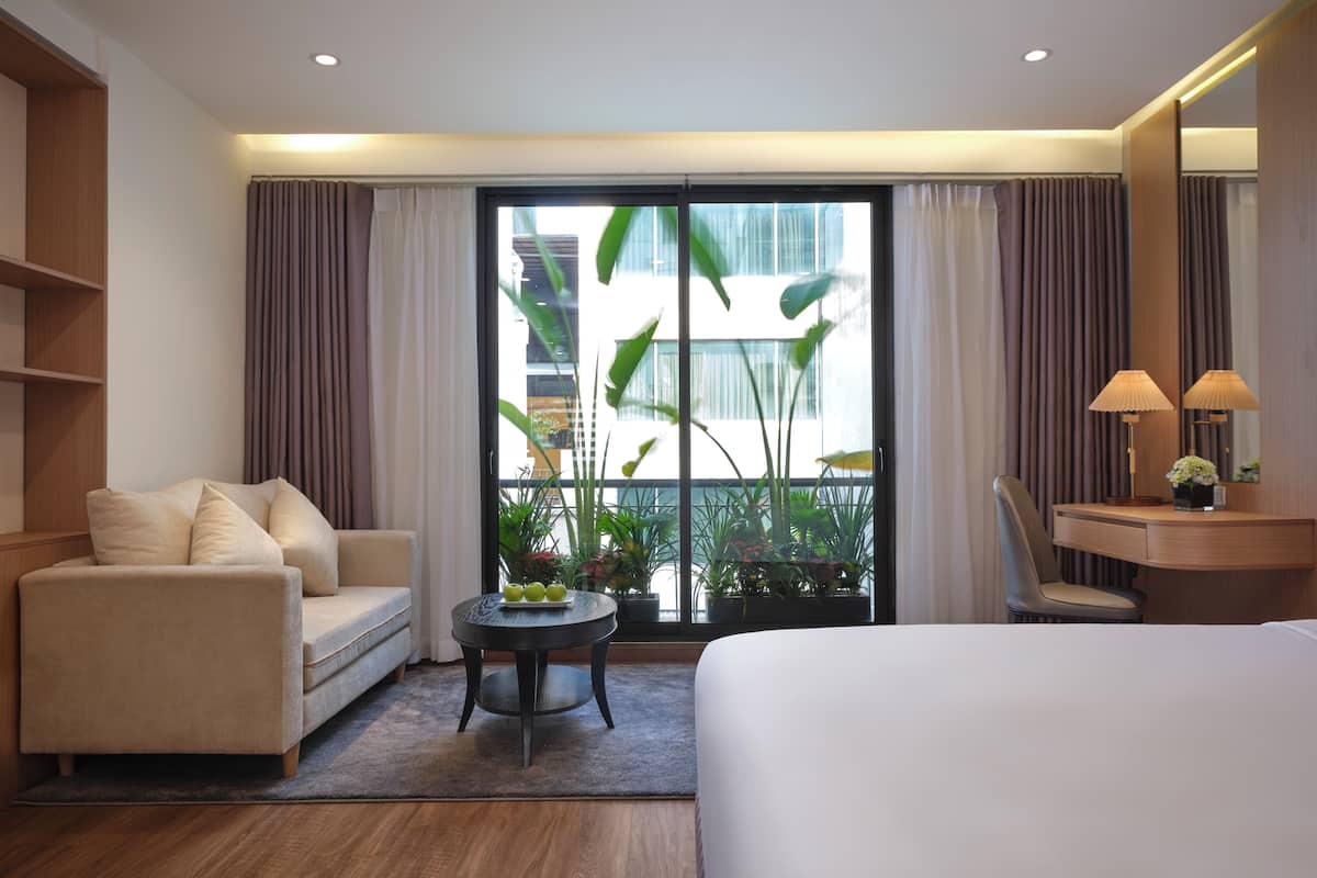 A hotel room with a bed, a sofa, a desk with a lamp, a small table with fruit, and a view of greenery outside.