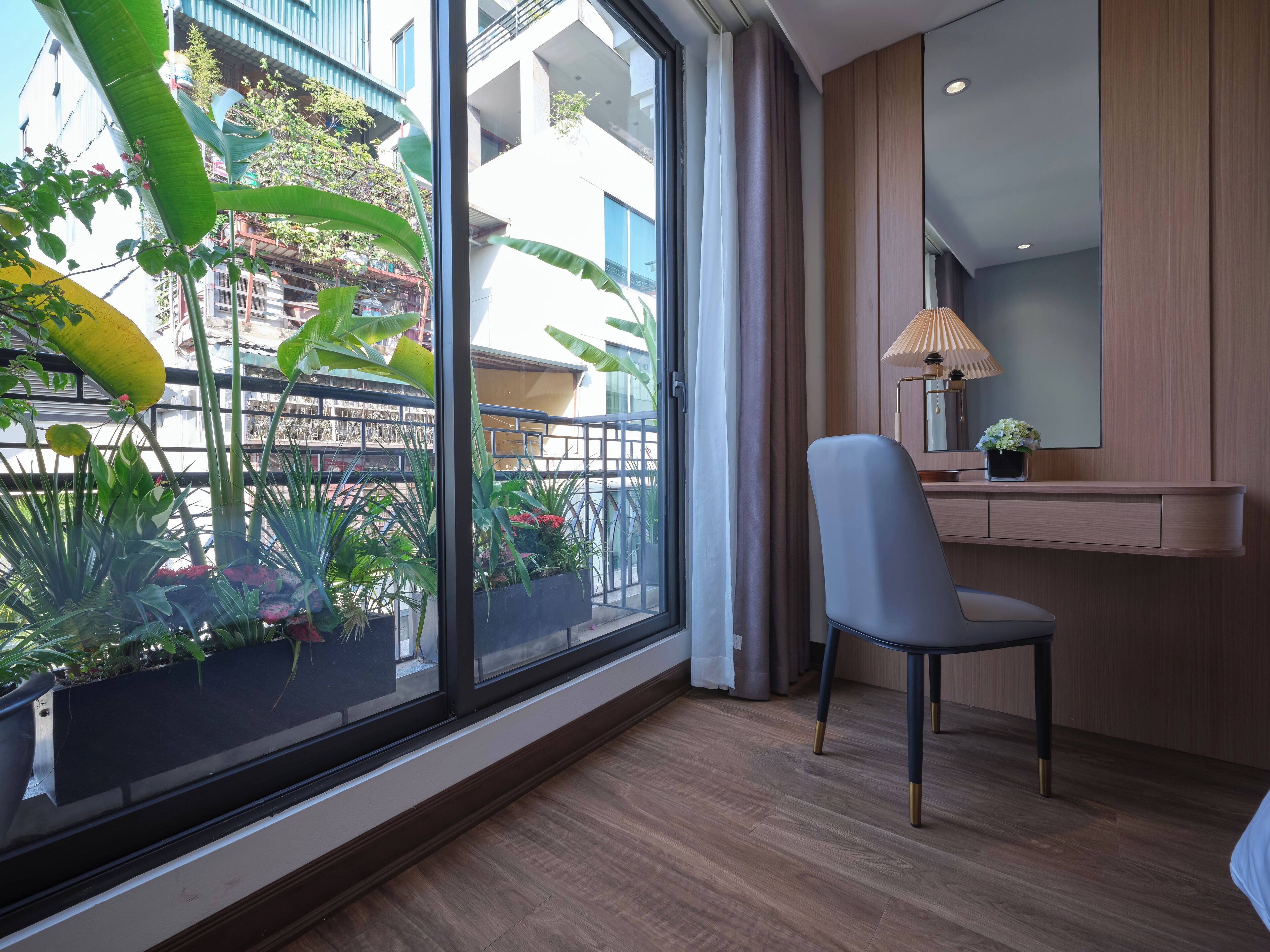 A modern hotel room with a large window, a chair, a bedside table with a lamp, and a mirror.