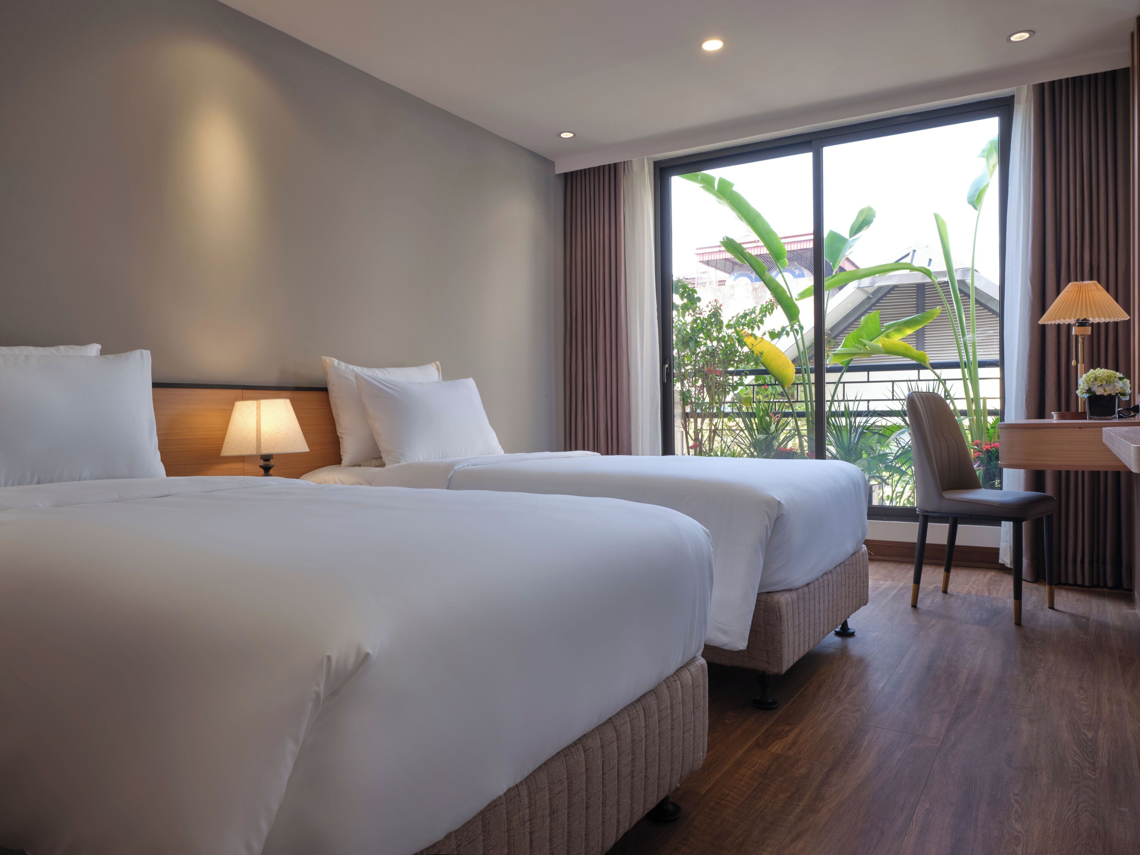A hotel room with two beds, a desk, a chair, and a large window with a view of greenery.