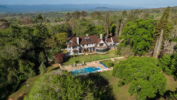 Una casa grande de dos pisos con piscina, rodeada de bosque denso y montañas al fondo.