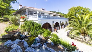 A two-story house with a balcony, a garden with a pond, and a pathway leading to the entrance.