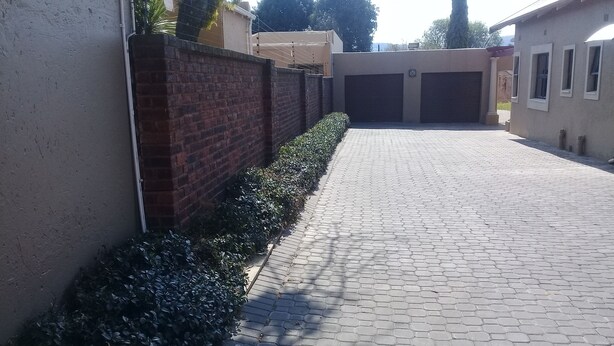 Une allée pavée menant à un garage, bordée d’un mur en briques et d’une haie.
