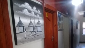 Un couloir doté d’un plancher en bois, une porte avec un interrupteur et un tableau accroché au mur représentant une scène avec des huttes et des personnages.