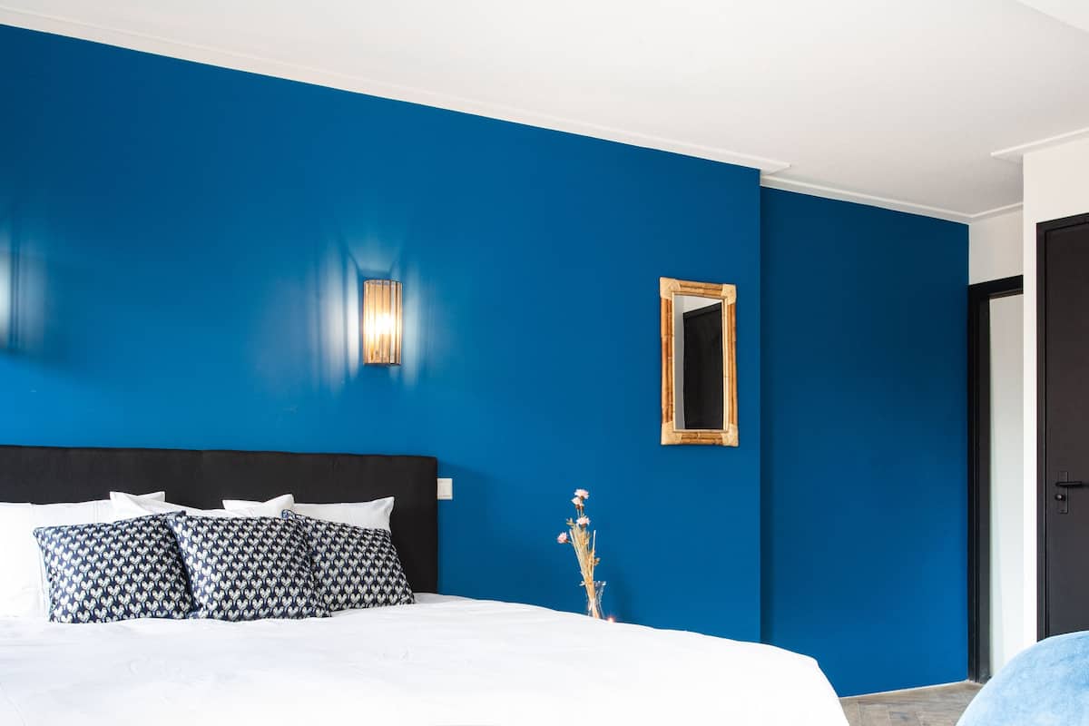 Un dormitorio con pared azul, cabecero negro, ropa de cama blanca y dos lámparas fijadas a la pared.