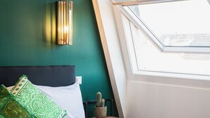 Una cama bien hecha con sábanas blancas y dos toallas dobladas, un cactus en una mesita de noche, y una pared verde con una lámpara fijada a la pared.