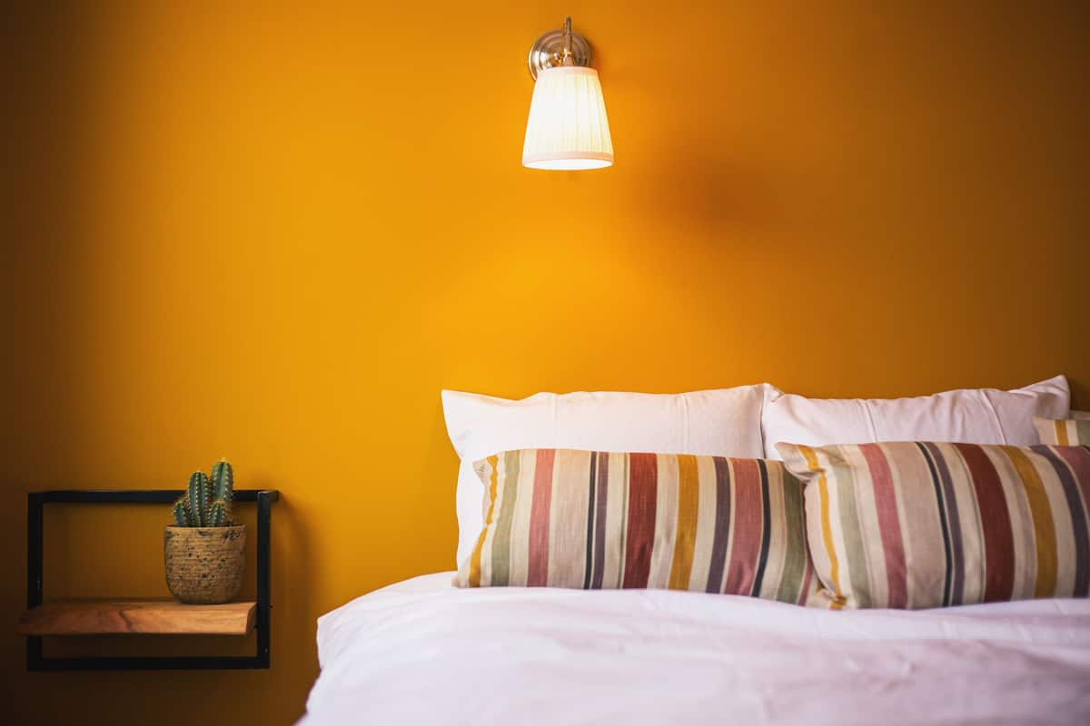 Una cama con ropa de cama blanca, almohadas a rayas y un pequeño estante con un cactus.