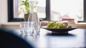 Una mesa con una jarra de vidrio transparente, cuatro vasos y un plato de uvas verdes.