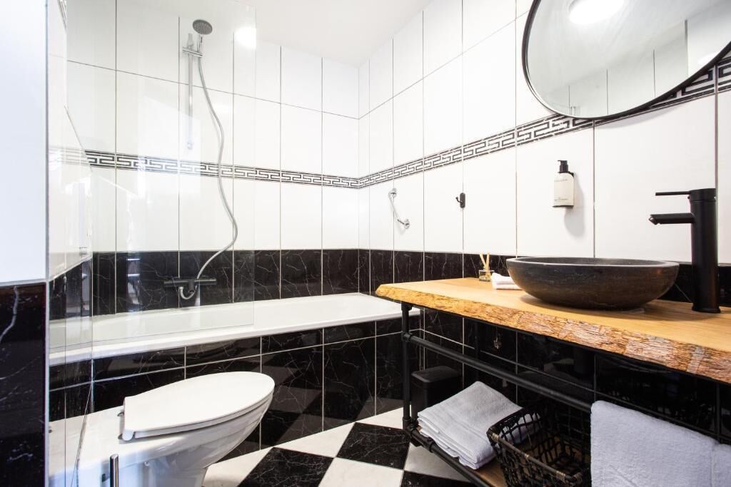 A modern bathroom with a black and white checkered floor, a white toilet, a large mirror, a wooden vanity with a black sink, and a black bathtub.