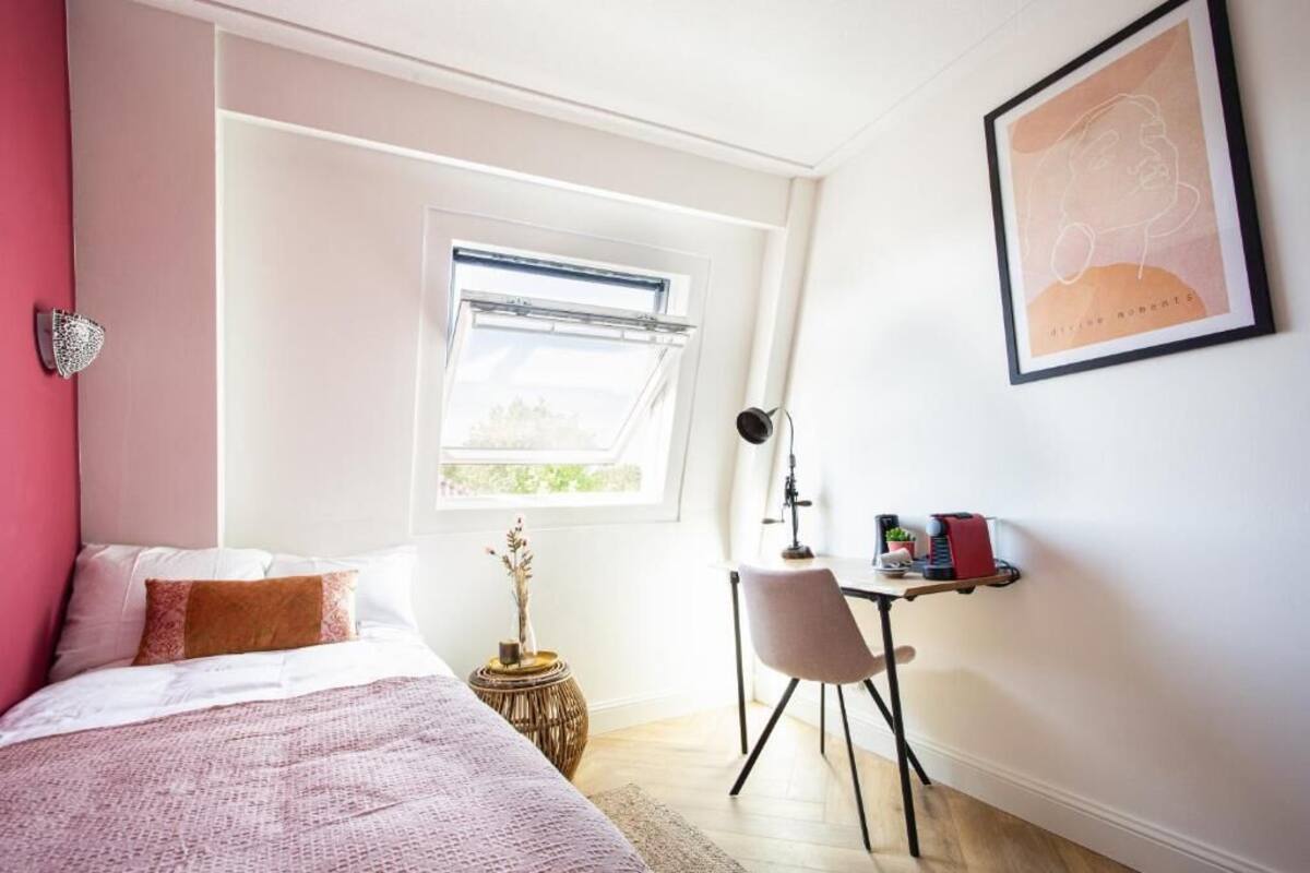 Un dormitorio con cama, escritorio, silla, ventana y lámpara de pared.