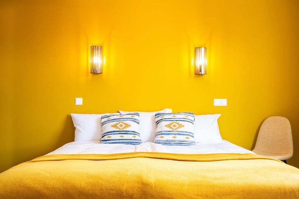 Habitación de hotel con pared amarilla, dos luces fijadas a la pared, cama con ropa blanca y almohadas estampadas, y una sola silla.