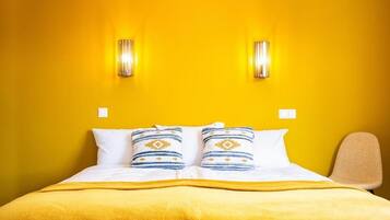 Habitación de hotel con pared amarilla, dos luces fijadas a la pared, cama con ropa blanca y almohadas estampadas, y una sola silla.