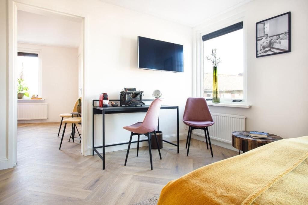 Ein modernes Zimmer mit Holzboden, einem Schreibtisch mit Computer, zwei Stühlen, einem Bett mit gelber Decke, einem Wandfernseher und einem Fenster mit Blick.