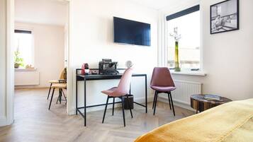 Ein modernes Zimmer mit Holzboden, einem Schreibtisch mit Computer, zwei Stühlen, einem Bett mit gelber Decke, einem Wandfernseher und einem Fenster mit Blick.