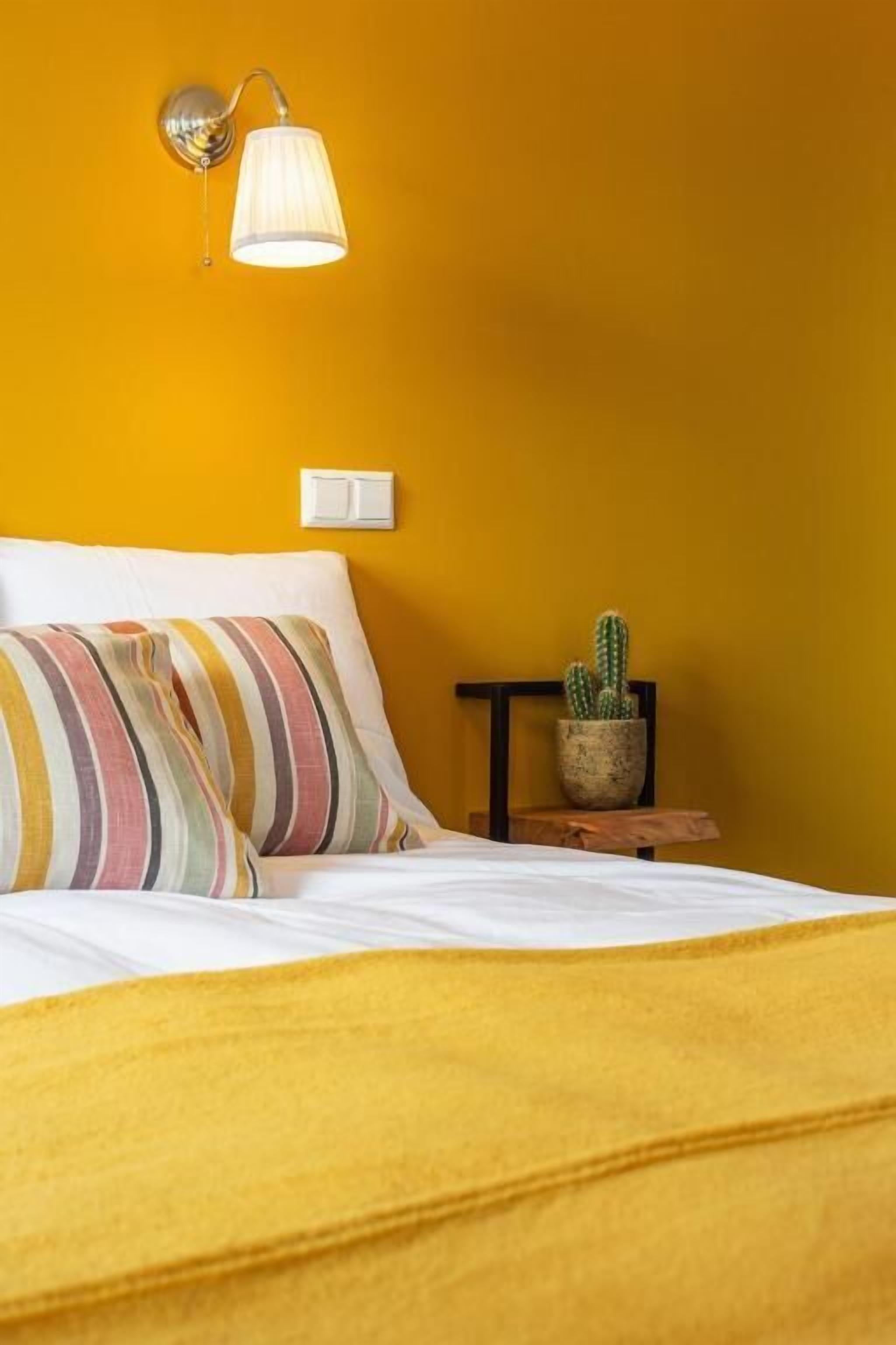 Una cama con almohadas a rayas, una lámpara fijada a la pared, un cactus sobre una mesita de madera y un interruptor de luz en la pared.