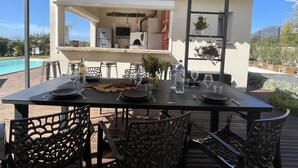 An outdoor dining area with a table set for two, surrounded by chairs, and a view of a pool and mountains in the background. - Magnificent new villa between sea and mountains, between Porticcio and Ajaccio (cauro)