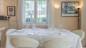 A dining area with a table set for four, white chairs, and a window with curtains. - Oliva Nova Golf Yellow House (OLIVA)