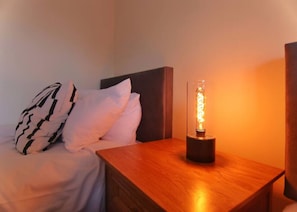 Un lit avec une literie blanche et des oreillers à motifs noir et blanc, une table de chevet en bois avec une lampe moderne, et un mur sobre en arrière-plan.
