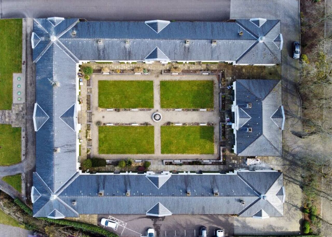 An aerial view of a parking lot with multiple parking spaces, a central roundabout, and surrounding green areas.