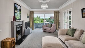 A living room with a fireplace, a television, a sofa, and a chair. There is a balcony with a view of greenery. - 12b Crest Rd · Escape Ordinary in Enniskillen (Enniskillen)