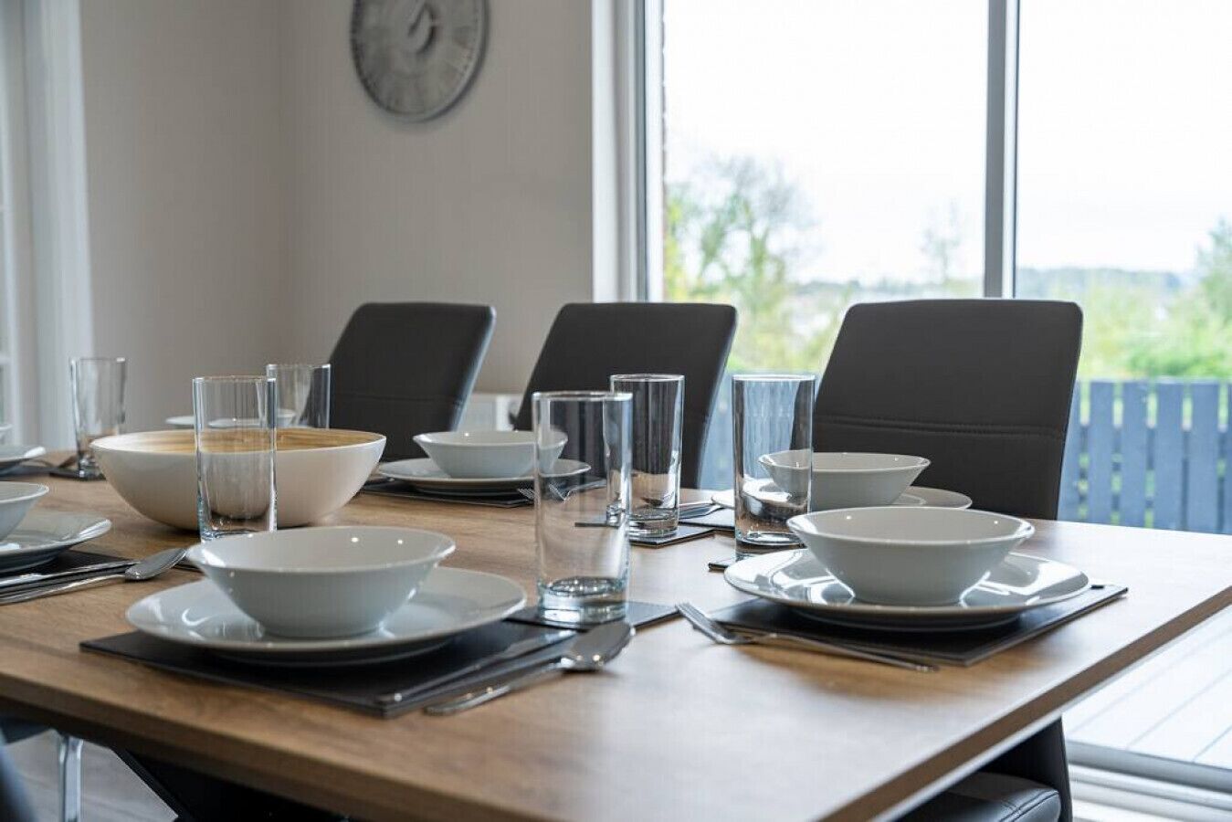 A dining table set with white plates, bowls, and clear glasses, with a clock on the wall and a view of greenery outside.