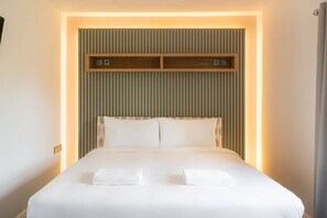 Una cama bien hecha con sábanas blancas y dos almohadas, un estante de madera encima y una pared con listones verticales.