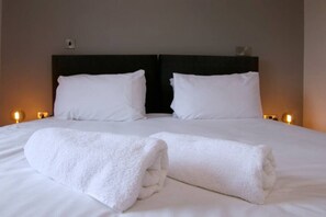 Una cama bien hecha con sábanas blancas y dos toallas enrolladas, cabecera oscura y lámparas de noche.