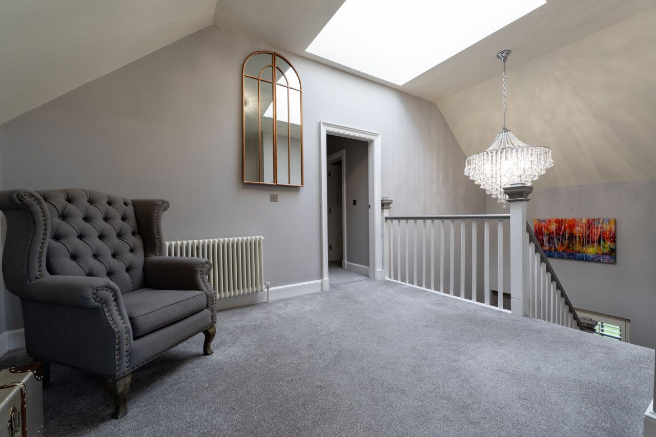 Une pièce spacieuse avec un plafond haut, une grande fenêtre, un fauteuil imposant, un radiateur, un escalier et un lustre.