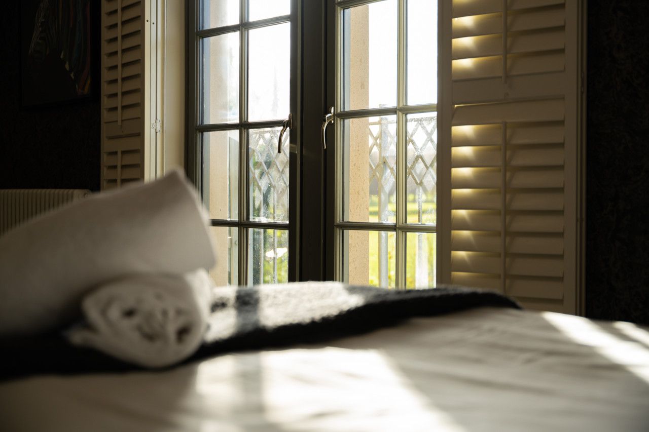 A bed with a rolled towel, a window with blinds, and a view of greenery outside.